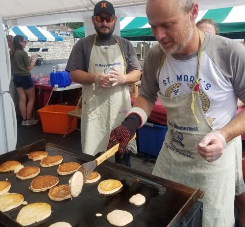 Two men in aprons cook pancakes on a griddle at an outdoor event, with tents and people in the background.