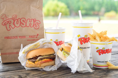 A takeout bag from Rush's with two burgers, fries, and drinks on a wooden table outdoors.