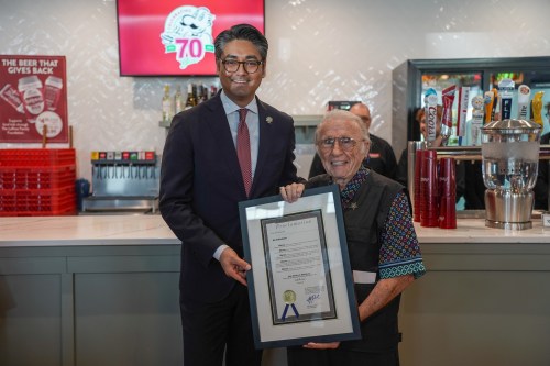A man in a suit stands next to an elderly man holding a framed proclamation in a bar setting.