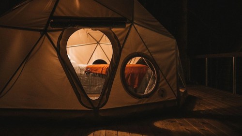 A cozy tent at night, illuminated from within, with a bed and warm blankets visible through circular windows.