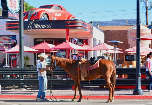 A man in a cowboy hat holds a horse near a retro diner with a red car on the roof and outdoor seating.