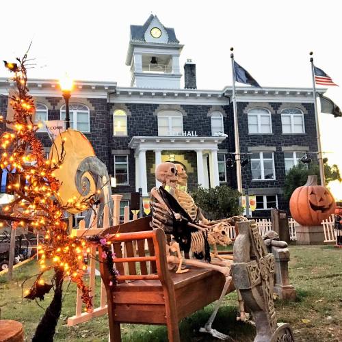A skeleton sits on a bench in front of a historic building, decorated for Halloween with pumpkins and lights.