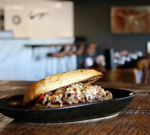 A close-up of a hearty sandwich on a black plate, featuring meat and toppings, set on a rustic wooden table.