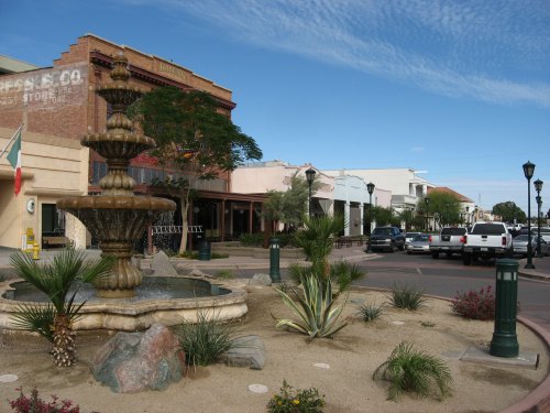 A street scene featuring a decorative fountain, palm trees, and shops under a clear blue sky.