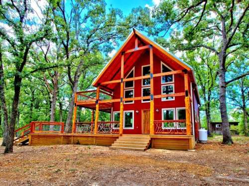 A red cabin with a steep roof, surrounded by trees, featuring a wooden deck and steps leading to the entrance.
