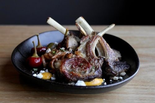 A black plate with grilled lamb chops, cherry peppers, and garnishes, served on a wooden table.