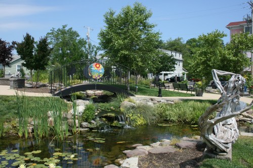 A serene park scene featuring a pond, a decorative bridge, lush greenery, and a sculpture near a walking path.