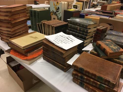 A table covered with stacks of various old books and a sign, surrounded by more books in the background.
