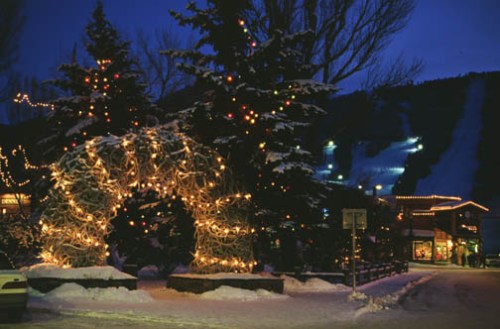 A snowy scene at night featuring trees adorned with colorful lights and a festive archway, with a ski slope in the background.