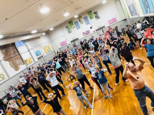 A large group of people dancing together in a spacious gym, with colorful decorations on the walls.