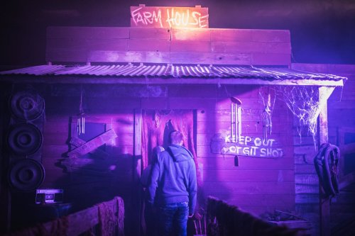 A dimly lit farmhouse with a "Keep Out or Get Shot" sign, surrounded by eerie decorations and purple lighting.