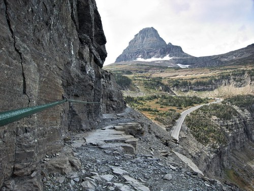 A rocky cliffside path with a metal railing, overlooking a valley and a distant mountain under a cloudy sky.