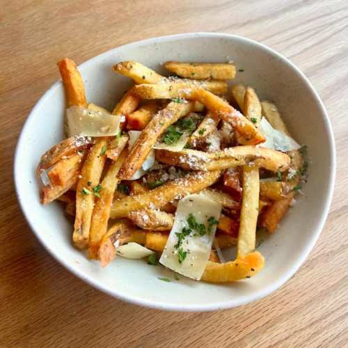 A bowl of golden-brown fries topped with grated cheese and herbs on a wooden surface.
