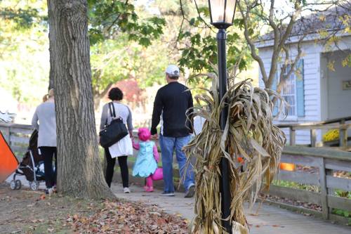 A family walks along a path, with a scarecrow decoration nearby and autumn foliage in the background.