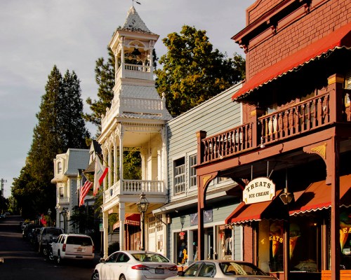 Historic buildings line a charming street, featuring a clock tower and shops, with trees in the background.