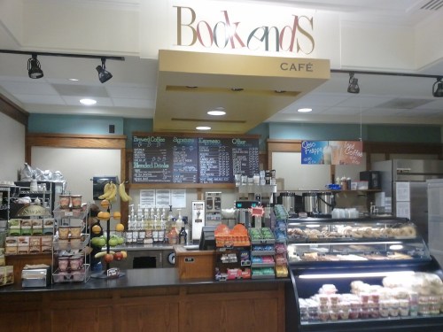 A café counter with a menu board, coffee machines, pastries, and fresh fruit displays. Cozy and inviting atmosphere.
