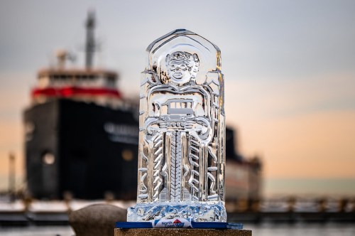 An ice sculpture of a figure stands in the foreground, with a large ship blurred in the background at sunset.