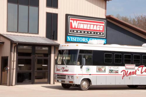 A white tour bus parked outside the Winnebago Industries Visitors Center, with a sign displaying the company name.