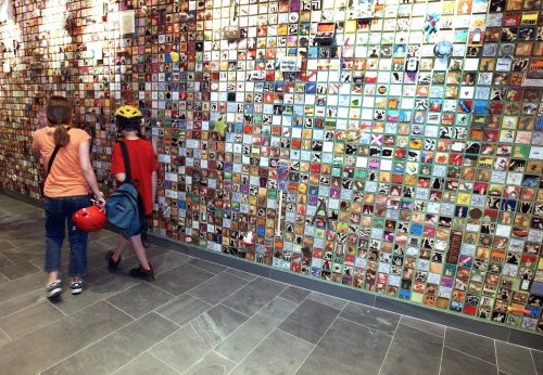 Two children walk past a colorful wall covered in small square art pieces, one wearing a helmet and the other a backpack.