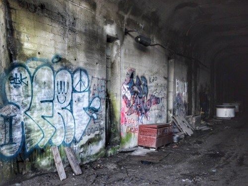 Graffiti-covered walls in a dimly lit, abandoned tunnel with debris scattered on the ground.