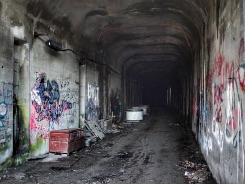 A dimly lit, graffiti-covered tunnel with debris and empty containers along the walls.