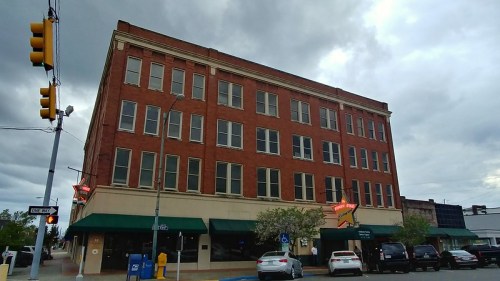 Historic red brick building with large windows, green awnings, and parked cars under a cloudy sky.