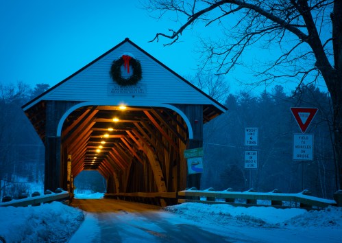 A snowy scene featuring a covered bridge adorned with a wreath, illuminated in the evening blue light.