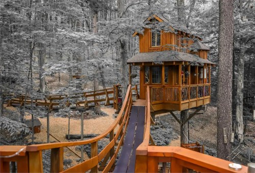 A wooden treehouse nestled in a forest, connected by a bridge, surrounded by trees in a serene, monochrome setting.