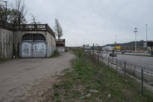 A gravel path beside a highway, with a concrete wall and graffiti on one side, and traffic in the distance.