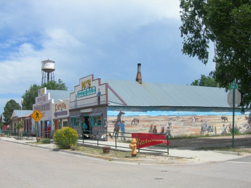 A colorful western-style building with a mural, featuring a bar and shops, set against a blue sky and water tower.