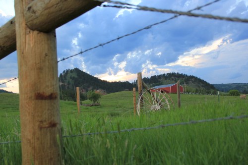 A scenic landscape featuring green fields, a wooden fence, a wagon wheel, and a red barn against a cloudy sky.