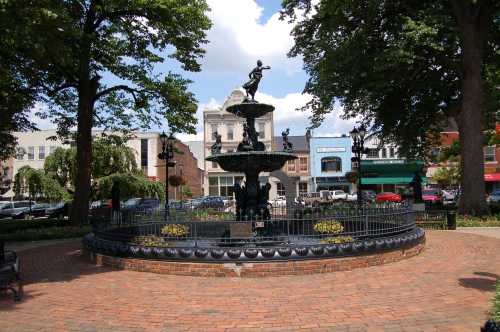 A decorative fountain surrounded by trees and shops, with a blue sky and clouds in the background.