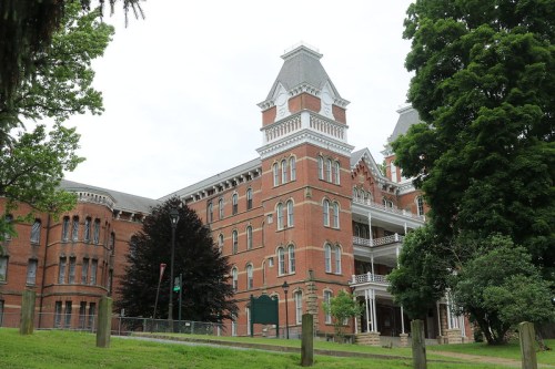 Historic red brick building with a tall tower, surrounded by trees and a grassy area, under a cloudy sky.