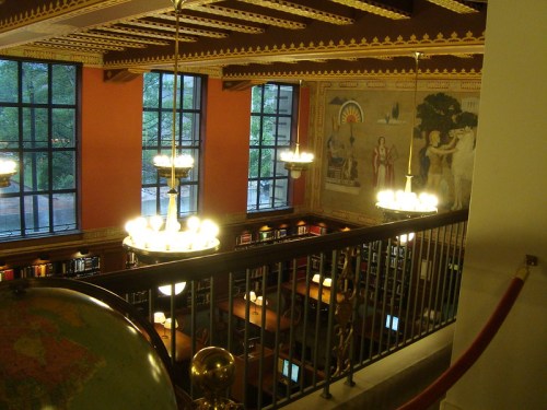 A view of a library interior with large windows, decorative murals, and wooden tables illuminated by chandeliers.