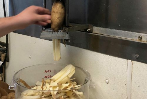 A hand operates a potato cutter, with freshly cut fries falling into a container below.