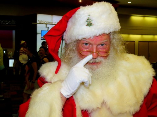 A jolly Santa Claus with a white beard and red suit playfully touching his nose, surrounded by festive decorations.