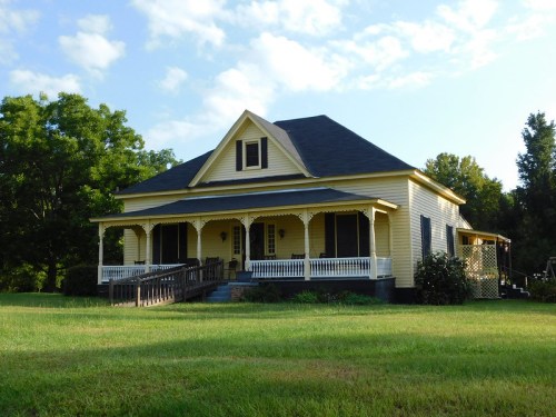 A charming yellow house with a porch, surrounded by green grass and trees under a blue sky.