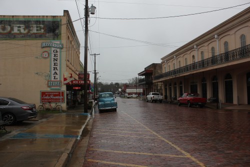 A rainy street scene featuring a general store, parked cars, and historic buildings with brick pavement.