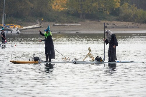 Two people in costumes stand on paddleboards in a lake, one with a witch hat and the other with long gray hair, accompanied by a skeleton.