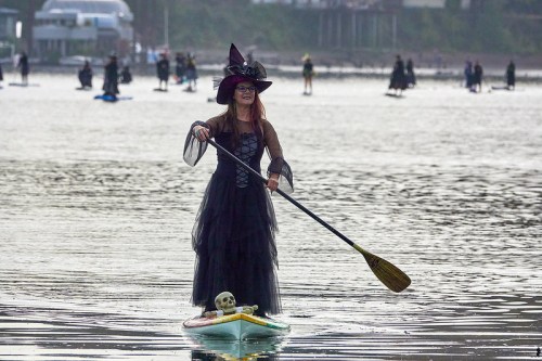 A woman in a witch costume paddles on a stand-up board, with a skeleton beside her, in a calm water setting.