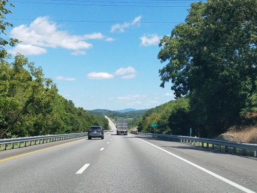 A highway stretches into the distance, flanked by trees and mountains under a clear blue sky with fluffy clouds.