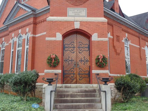 Historic red brick library with decorative wooden doors, wreaths on either side, and stone steps leading up.