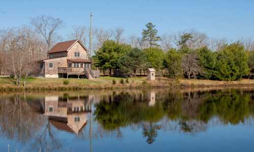 A serene house by a pond, surrounded by trees, reflecting in the calm water under a clear blue sky.