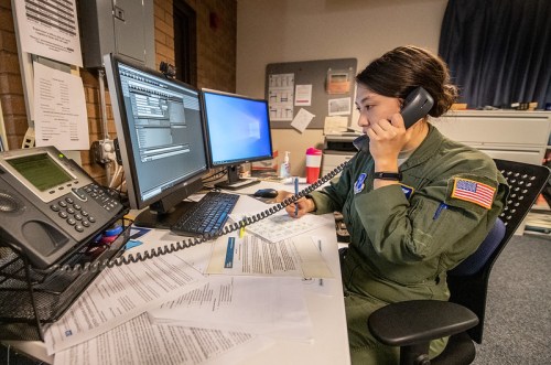 A service member in a flight suit talks on the phone while working at a desk with multiple computer screens and documents.
