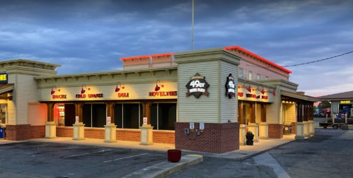 Exterior of a restaurant with a lit sign, featuring a covered patio and a parking lot under a cloudy sky.