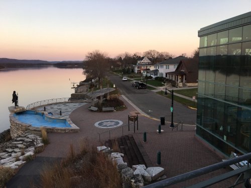 A serene riverside view at dusk, featuring a walkway, homes, and a modern building beside the water.