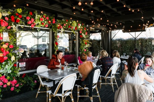A lively outdoor dining area adorned with flowers, featuring patrons enjoying drinks and conversation under string lights.