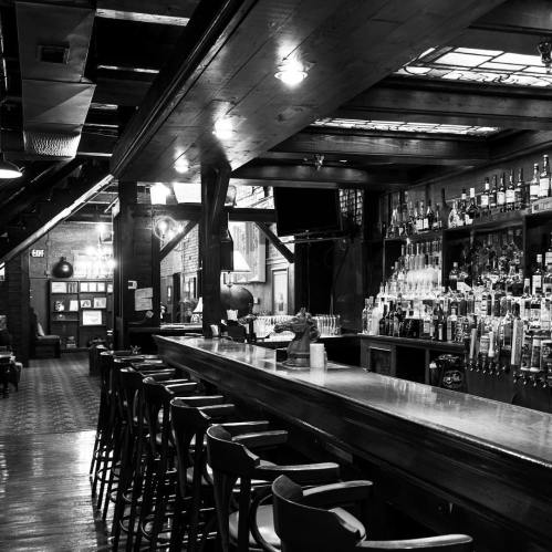 A dimly lit bar with a long wooden counter, high stools, and shelves lined with various bottles.
