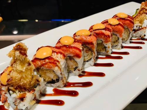 A plate of sushi rolls topped with spicy sauce and drizzled with sweet sauce, alongside a crispy tempura piece.