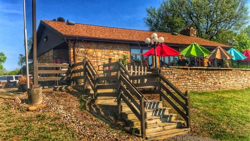 A stone building with a sloped wooden staircase, colorful umbrellas, and outdoor seating on a sunny day.